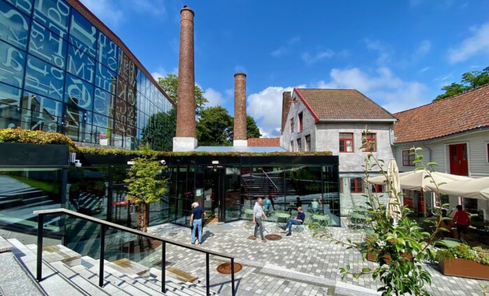 Uteområdet hos Iddis i Gamle Stavanger, med glassfasade, gamle pipeløp fra hermetikkfabrikken og hvite trehus. Søndagsåpne kafeer i Stavanger