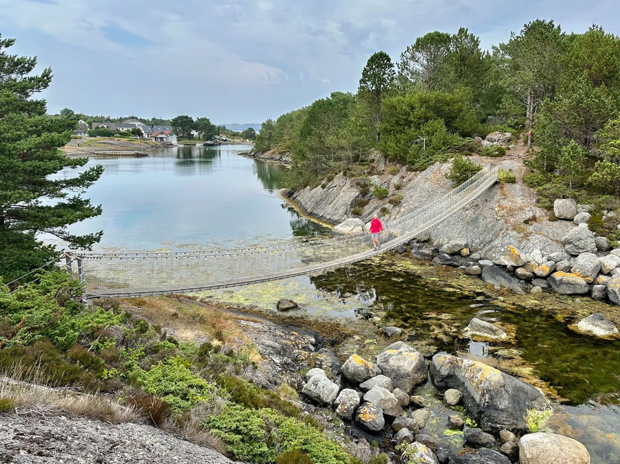 Person i rød jakke går over hengebrua på Vassøy, med sjø, svaberg og furutrær rundt.
